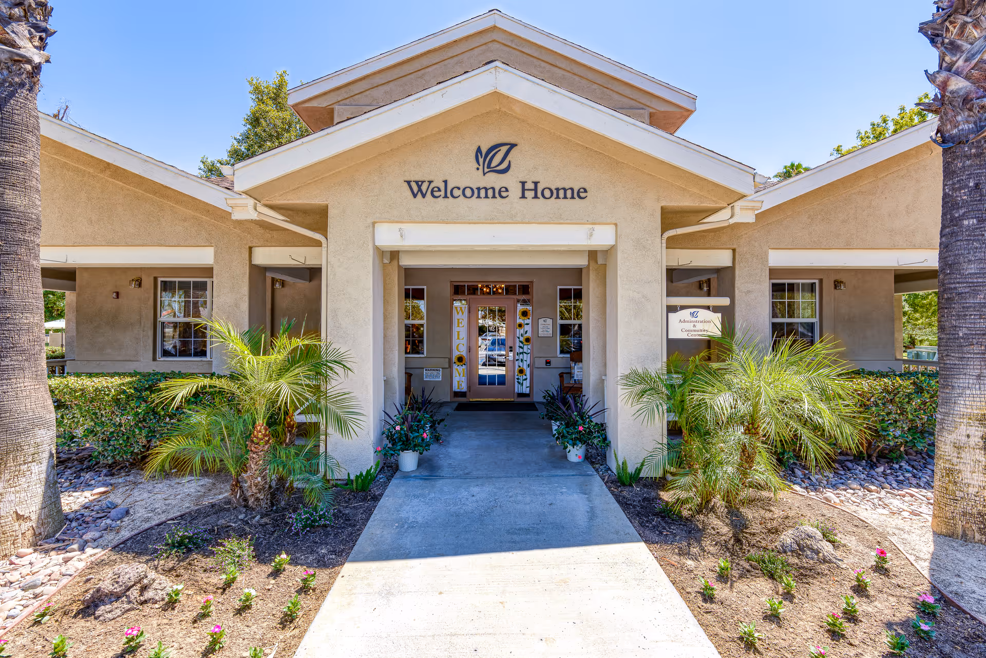 Front entrance of a beige senior living facility with a covered walkway, potted plants, and a 'Welcome Home' sign above the doorway.