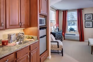 Studio-style apartment interior showing a kitchenette with wooden cabinets and sink on the left and a seating area with an armchair, dresser and window with red curtains on the right.