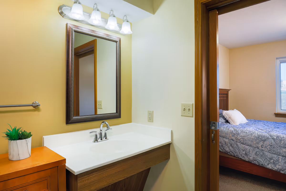 A bathroom vanity with a white countertop and a silver faucet, a framed mirror above it with four light fixtures. To the left, there is a small wooden cabinet with a potted plant on top. Through an open doorway, a bedroom is visible with a bed covered in a blue patterned bedspread and a window letting in natural light.