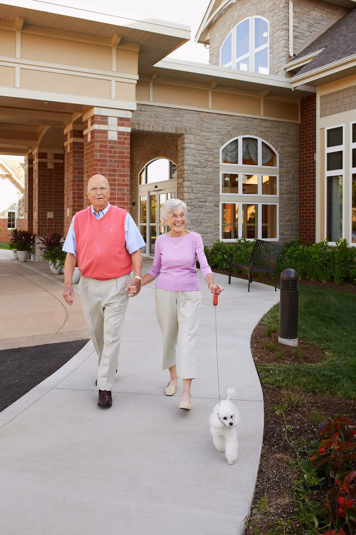 An elderly couple walking hand in hand on a curved sidewalk outside a senior living facility named Aberdeen Heights. The woman is holding a leash with a small white dog. The building behind them has brick and stone exterior with large windows and a covered entrance.