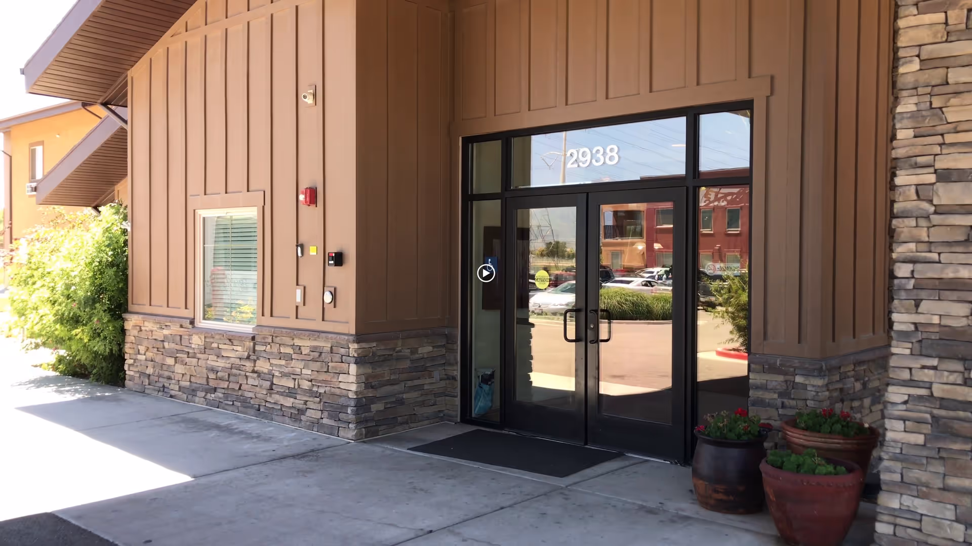 Entrance to a building with double glass doors numbered 2938, surrounded by brown paneling and stone accents. There are potted plants near the entrance and a sidewalk leading up to the doors.