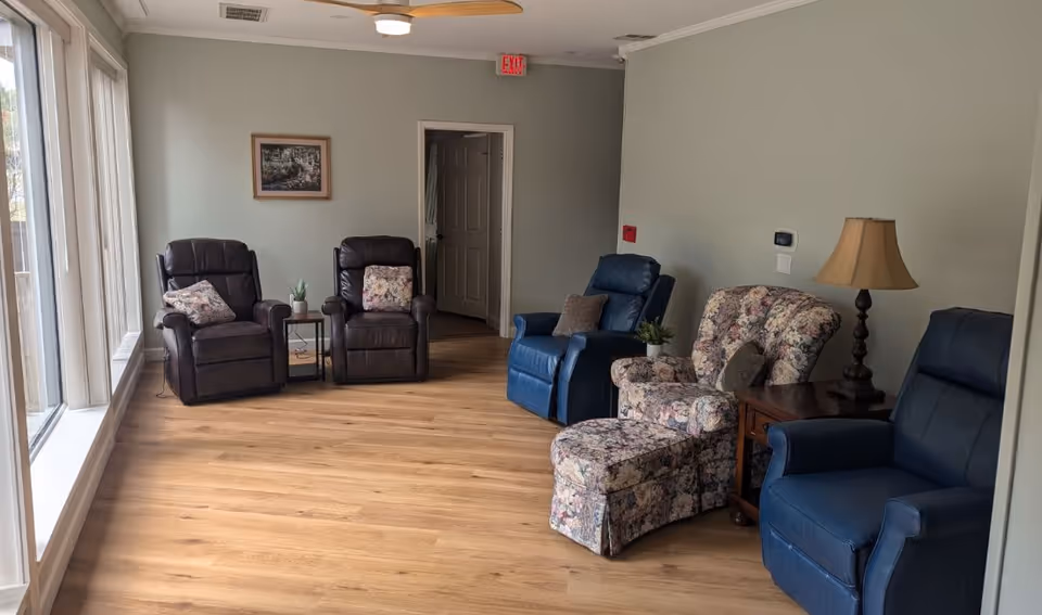 A cozy sitting area with five recliner chairs arranged along the walls. Two dark brown leather recliners with floral pillows are near large windows on the left. A floral patterned armchair with a matching ottoman, two blue recliners, a wooden side table with a lamp, and small potted plants complete the room. The floor is light wood, and the walls are painted light gray. A ceiling fan and an exit sign are visible on the ceiling and wall.
