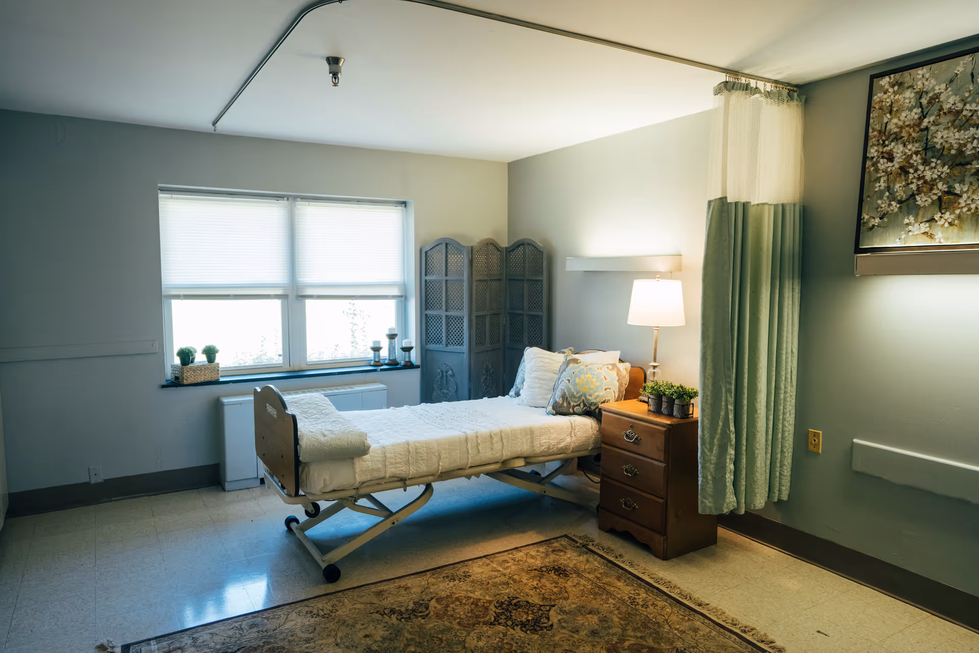 A simple, well-lit bedroom in a senior living facility with a single adjustable bed covered in white bedding. Next to the bed is a wooden nightstand with a lamp and small potted plants. A decorative folding screen is placed in the corner near a window with blinds. A curtain hangs from the ceiling, and a floral painting is mounted on the wall. The floor has a patterned rug.
