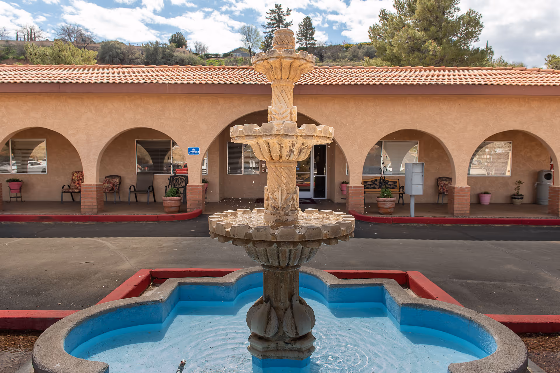 A stone water fountain with three tiers in front of a building with a tiled roof and arched walkways. There are chairs and potted plants under the arches, and a sign indicating van accessibility is visible on one of the columns.