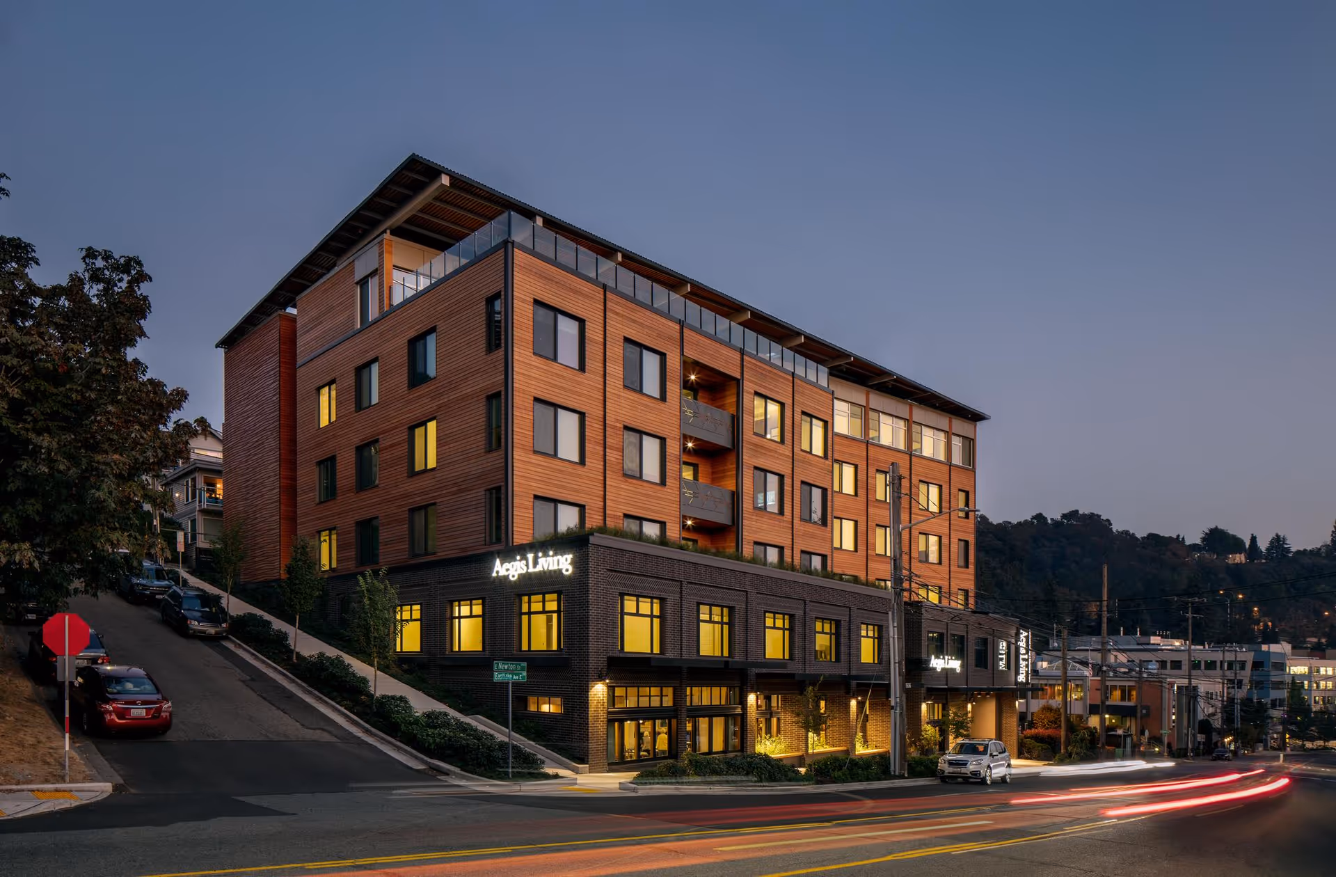 Exterior view of a modern multi-story building named Aegis Living Lake Union at dusk, with warm lights glowing from the windows and cars parked along the street.