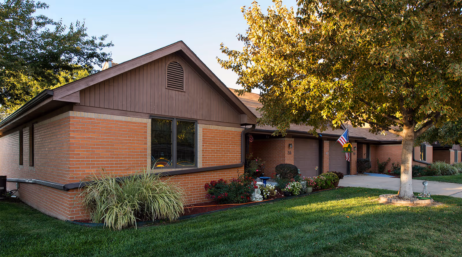 Exterior view of a single-story brick building with a brown roof and trim, surrounded by green grass, plants, and a large tree. There is an American flag and a birdhouse near the entrance, with a driveway leading to a garage door.