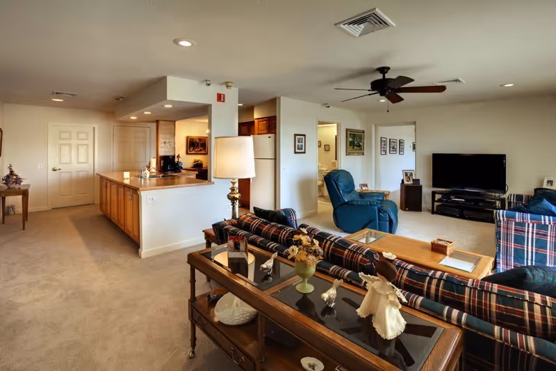 Open-plan living room with plaid sofas, a glass-topped console table, a recliner and TV, and a kitchen island visible in the background.