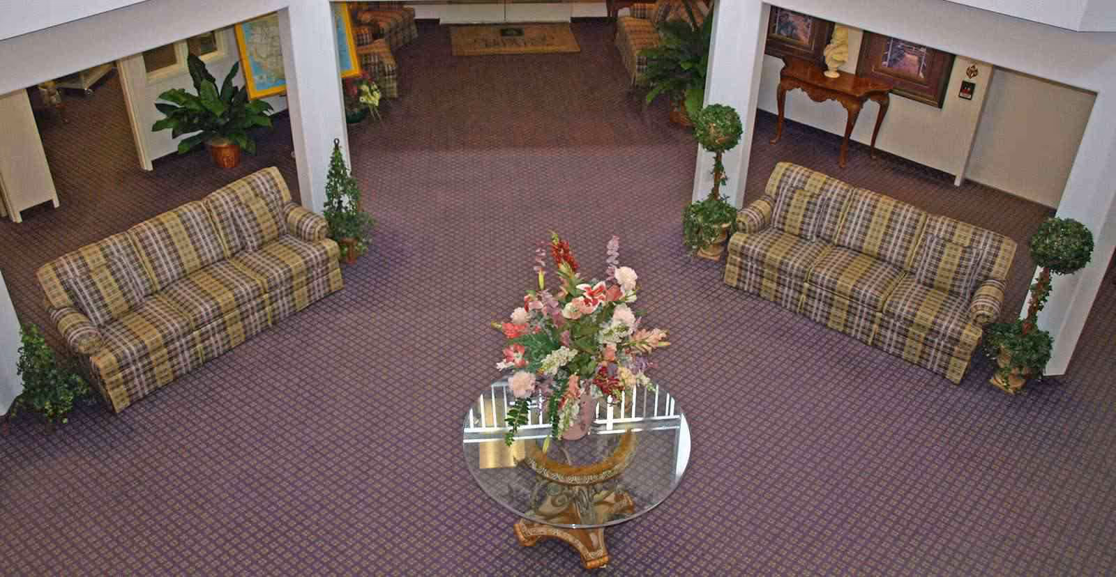 Interior view of a senior living facility lobby with two plaid sofas facing each other, a round glass table with a floral arrangement in the center, potted plants, and framed artwork on the walls.