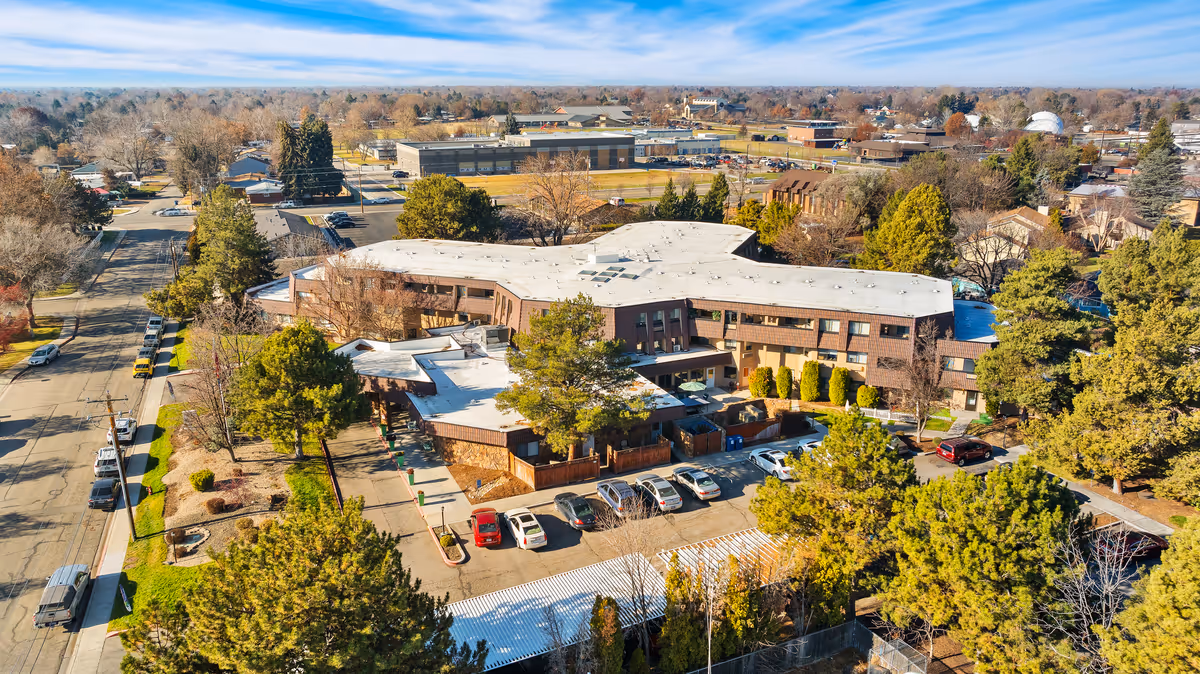 Aerial view of The Chateau de Boise, a senior living community building surrounded by trees and parking areas, with a neighborhood and other buildings in the background under a partly cloudy sky.