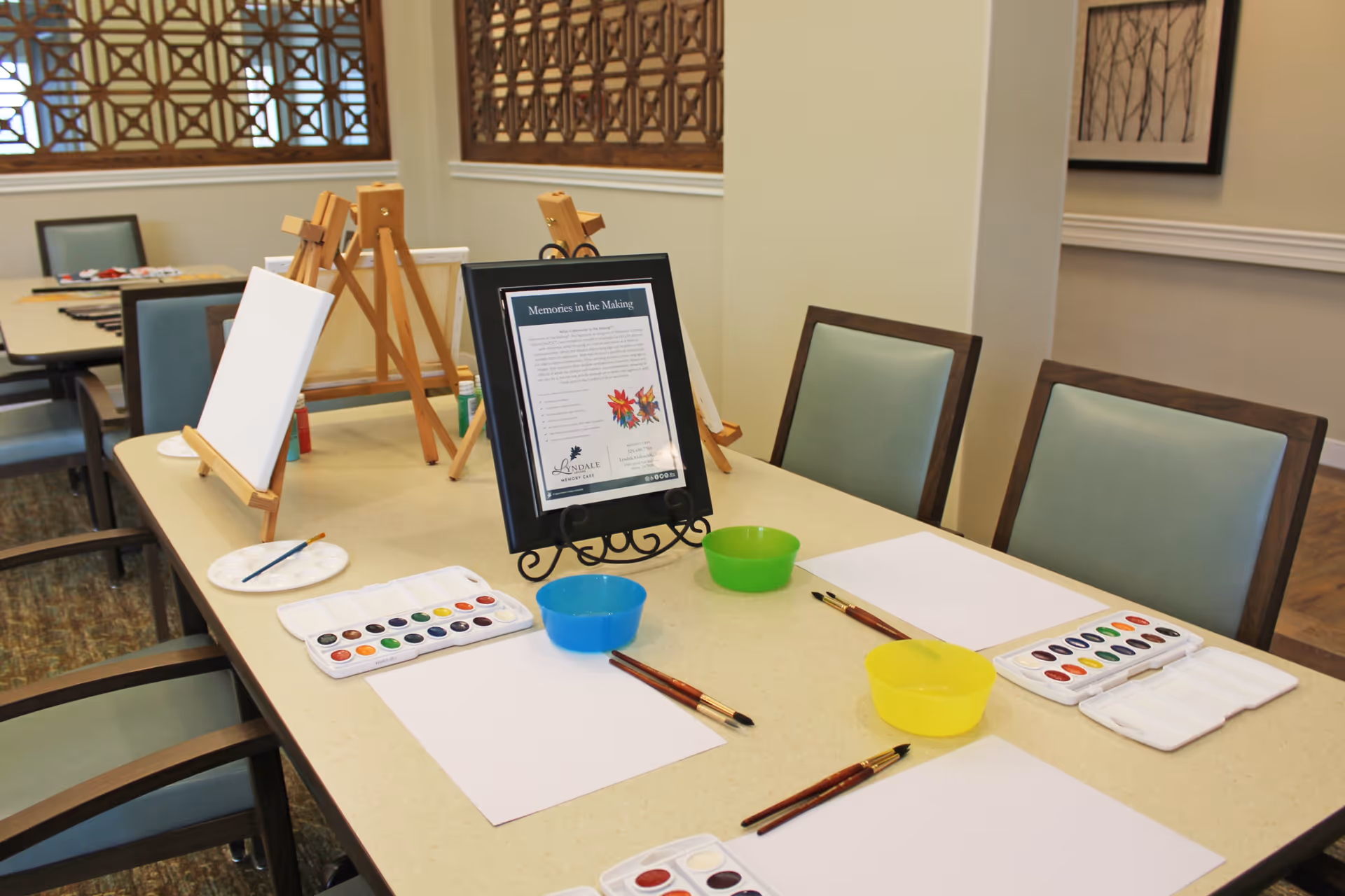 A table set up for a watercolor painting activity with blank canvases on easels, watercolor paint palettes, paintbrushes, bowls of water, and blank sheets of paper in a well-lit room with chairs around the table.