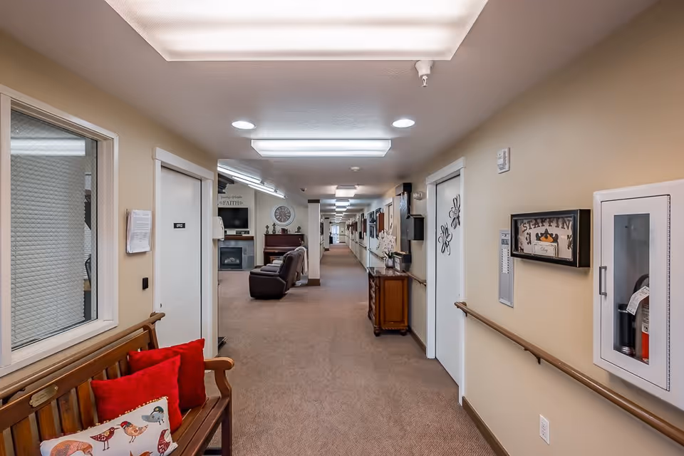 A long, well-lit hallway in a senior living facility with beige walls and carpeted floor. On the left side, there is a wooden bench with red and bird-patterned pillows. The hallway features handrails on both sides, several doors, a fire extinguisher cabinet, and a small wooden table with a flower arrangement. At the far end, there is a seating area with a piano, a clock on the wall, and a TV above a fireplace.