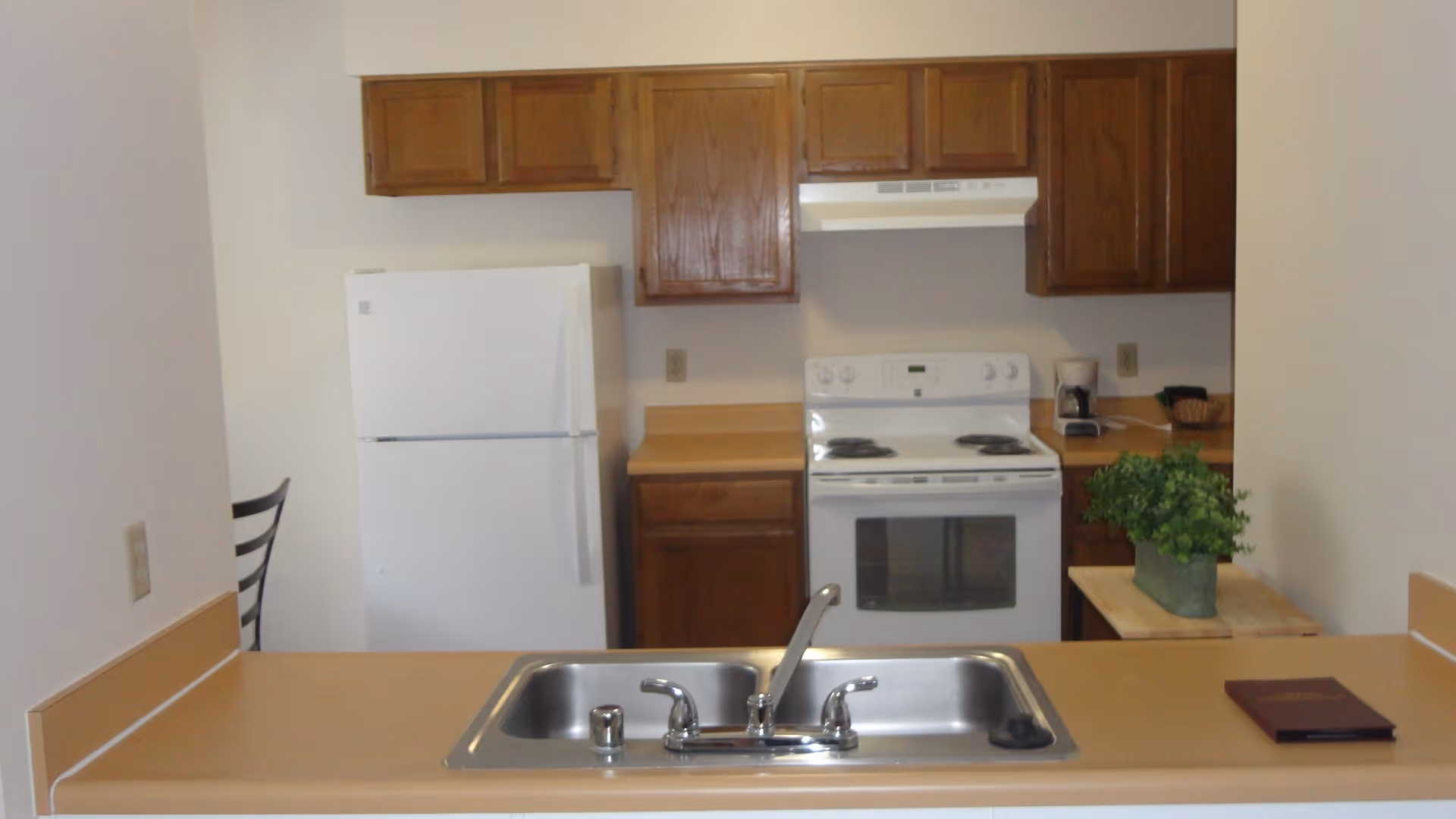 A kitchen area with a white refrigerator, white electric stove with oven, wooden cabinets, a double stainless steel sink with a faucet, a small potted plant on the counter, a coffee maker, and a chair partially visible behind the refrigerator.