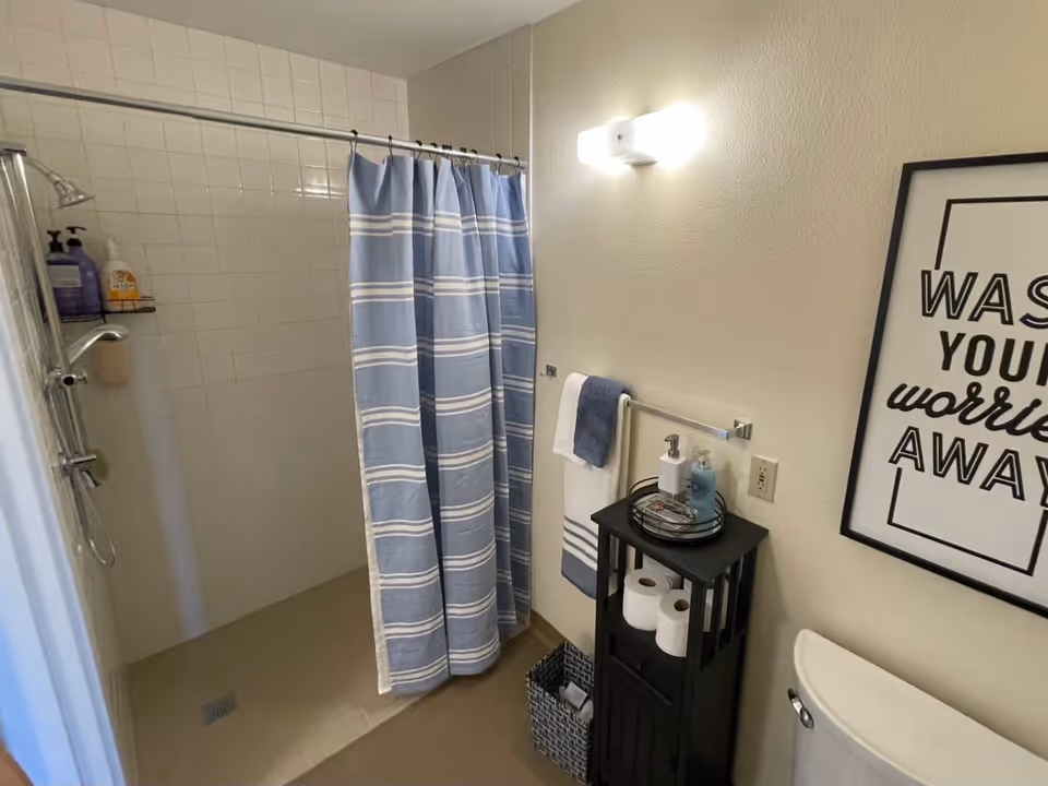 Bathroom with a walk-in shower featuring white tiled walls and a blue and white striped shower curtain. A black cabinet holds toilet paper rolls and hand soap, next to a white toilet. A towel rack with white and blue towels is mounted on the wall, which also has a framed sign that reads 'WASH YOUR worries AWAY'.