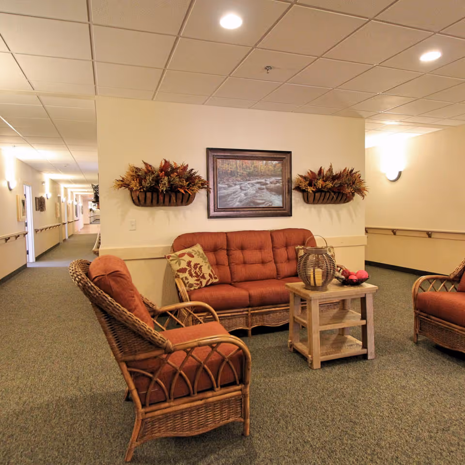 A cozy seating area in a senior living facility hallway featuring a wicker sofa and two matching chairs with rust-colored cushions. A small wooden table with a decorative lantern and bowl sits in the center. The wall behind the sofa has a framed picture of a forest stream and two wall-mounted planters with autumn foliage. The hallway is carpeted and well-lit with ceiling lights and wall sconces.