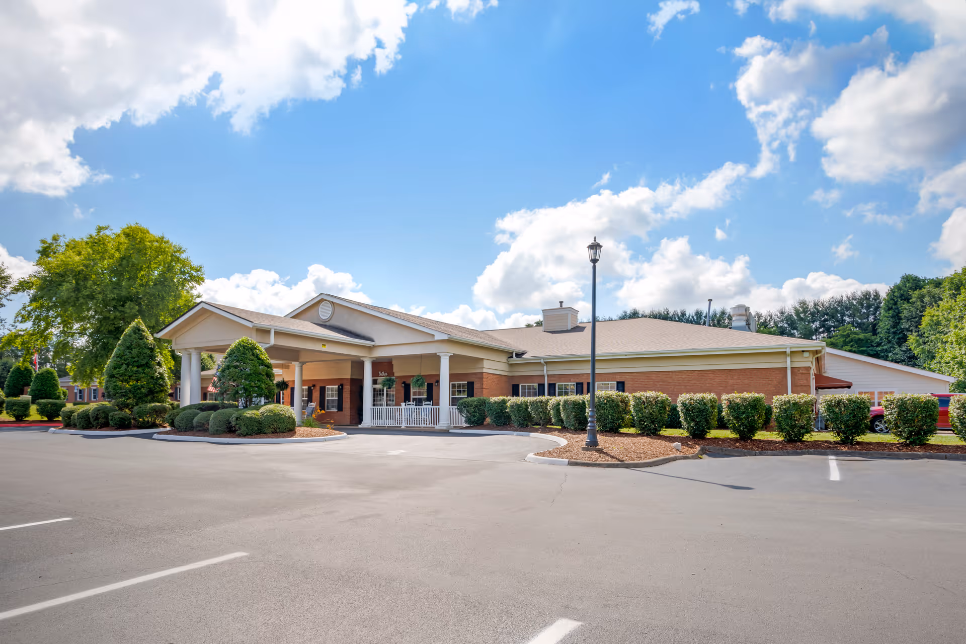 Exterior view of a single-story senior living facility building with a covered entrance, surrounded by neatly trimmed bushes and trees under a partly cloudy blue sky. The parking lot in front is mostly empty.