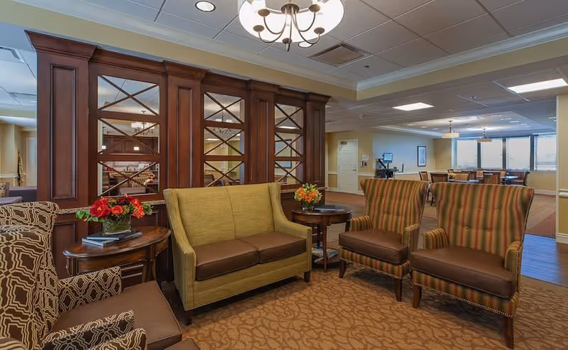 Comfortable lounge in a senior living facility with upholstered sofa and armchairs arranged around side tables and a wooden room divider leading to a dining area.