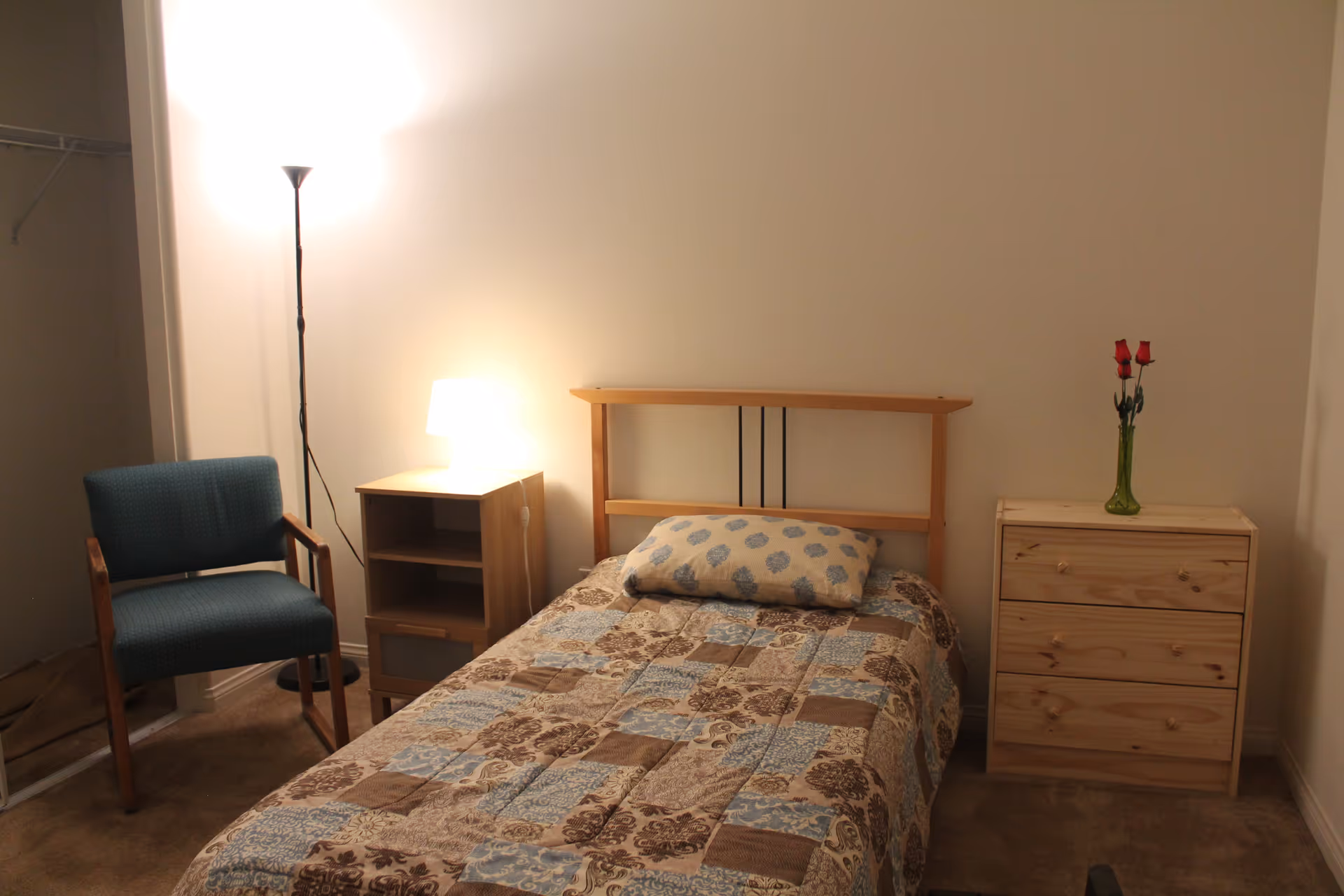 Small bedroom with a single bed, patterned bedspread, wooden headboard, nightstand, dresser with a vase of roses, an upholstered chair, and a floor lamp.