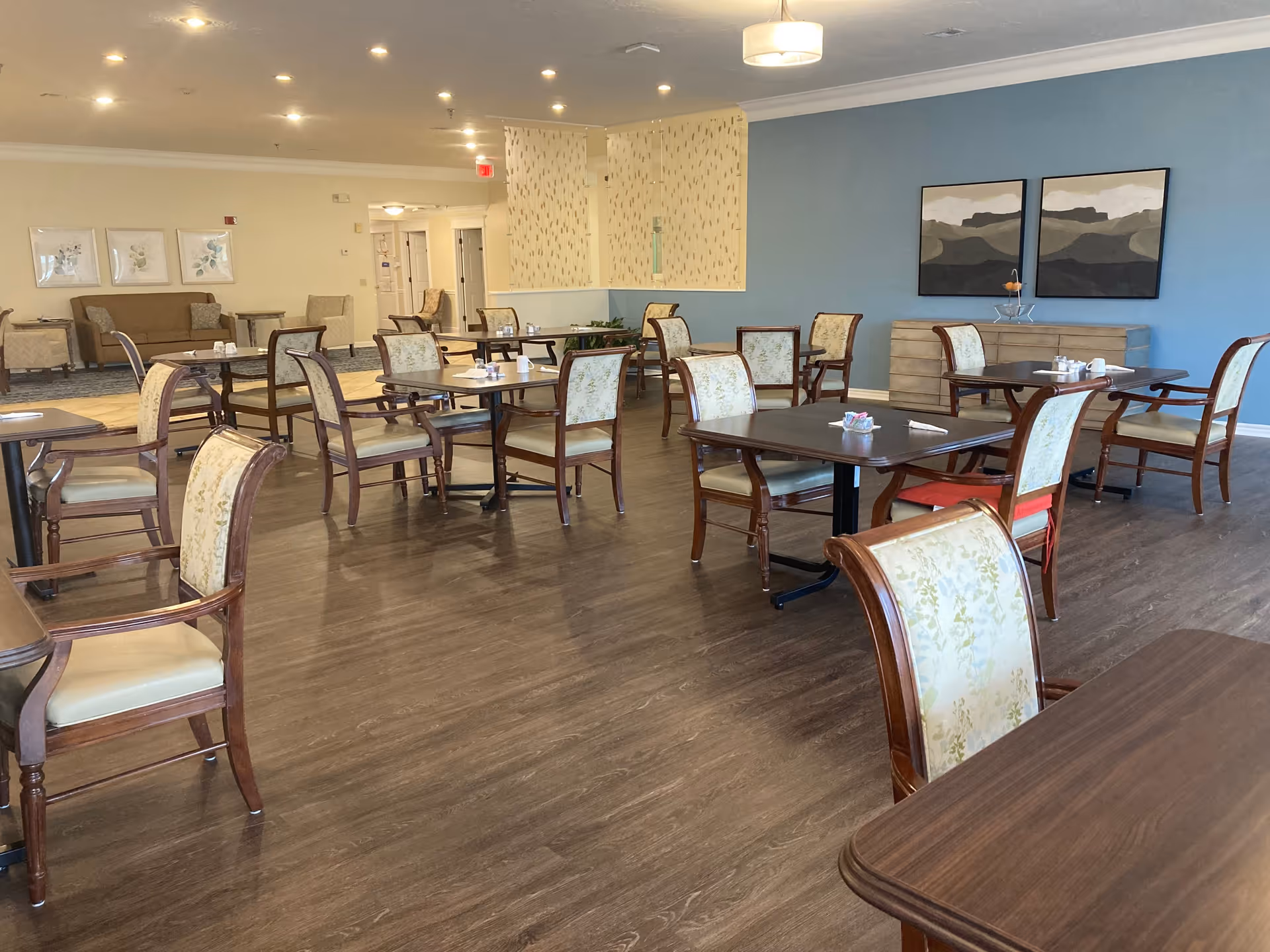 A spacious dining room with several wooden tables and chairs arranged neatly on a dark wood floor. The walls are painted light blue and beige, decorated with framed artwork and a sideboard. The room is well-lit with ceiling lights and a hanging lamp.