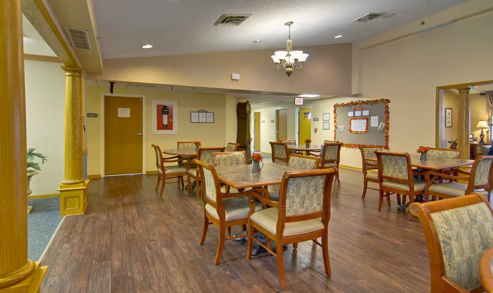 A senior living facility common dining area with multiple wooden tables and chairs arranged neatly. Each table has a small vase with flowers. The room has wood flooring, beige walls, and a chandelier hanging from the ceiling. There is a bulletin board decorated with autumn-themed garlands on one wall and a hallway with doors in the background.