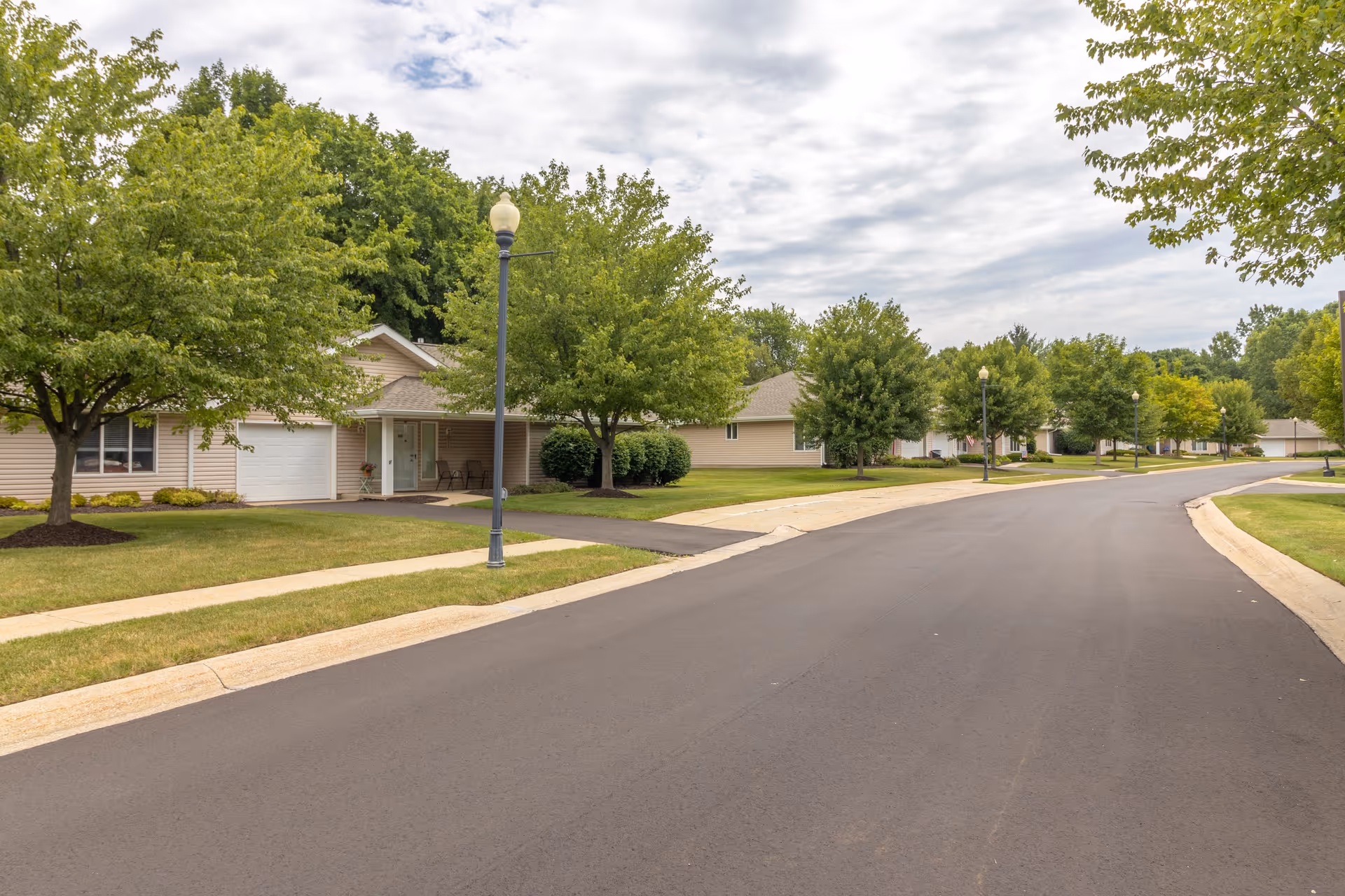 A quiet residential street in a senior living community with single-story beige houses, green lawns, trees, and street lamps under a partly cloudy sky.