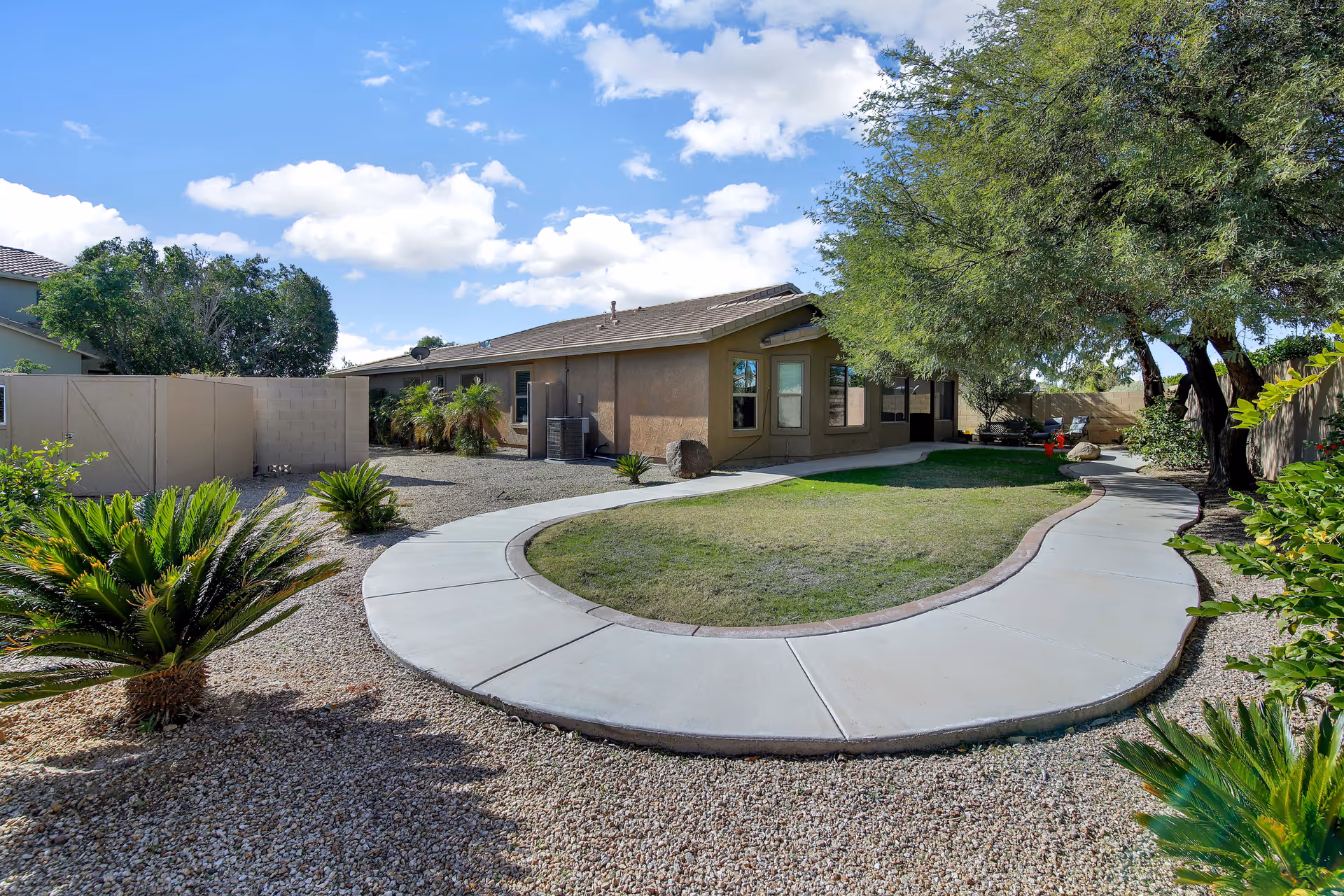 A curved concrete walkway surrounds a small grassy area in a backyard with desert landscaping including small palm plants and gravel. There are trees providing shade on the right side, and a single-story building with beige walls and multiple windows is visible in the background under a partly cloudy blue sky.