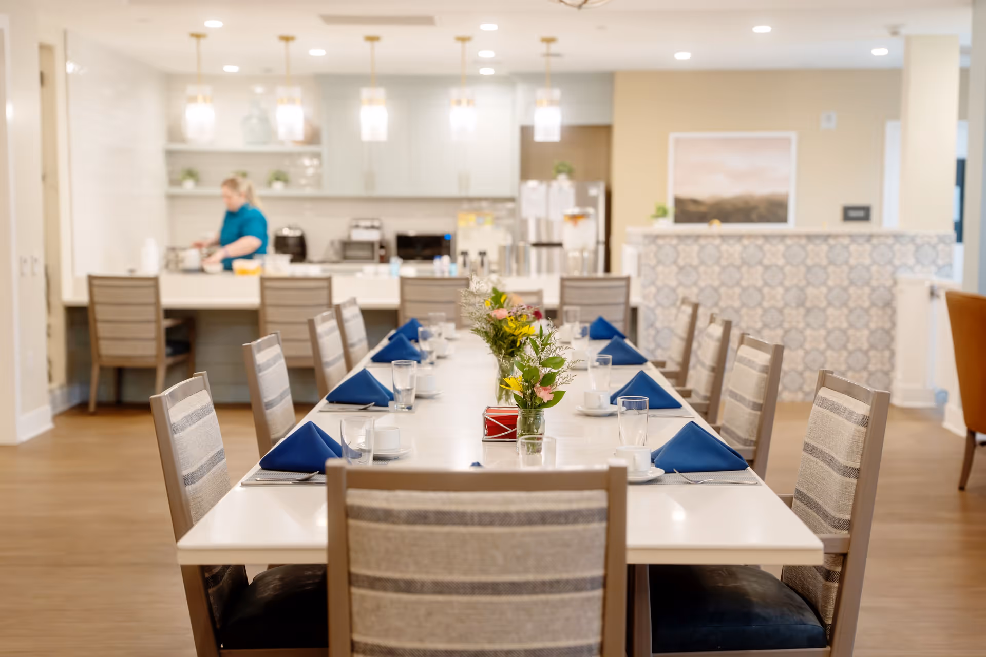 A long dining table set with blue folded napkins, glasses, cups, and small flower arrangements in a bright, modern dining room. In the background, a person is preparing food in a kitchen area with white cabinets and hanging pendant lights.