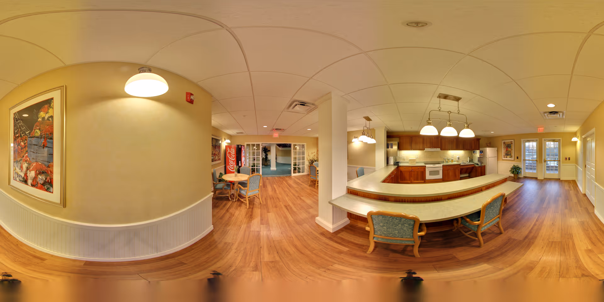 Panoramic view of a communal dining and kitchen area with a curved breakfast bar, tables, chairs, and wood floors.