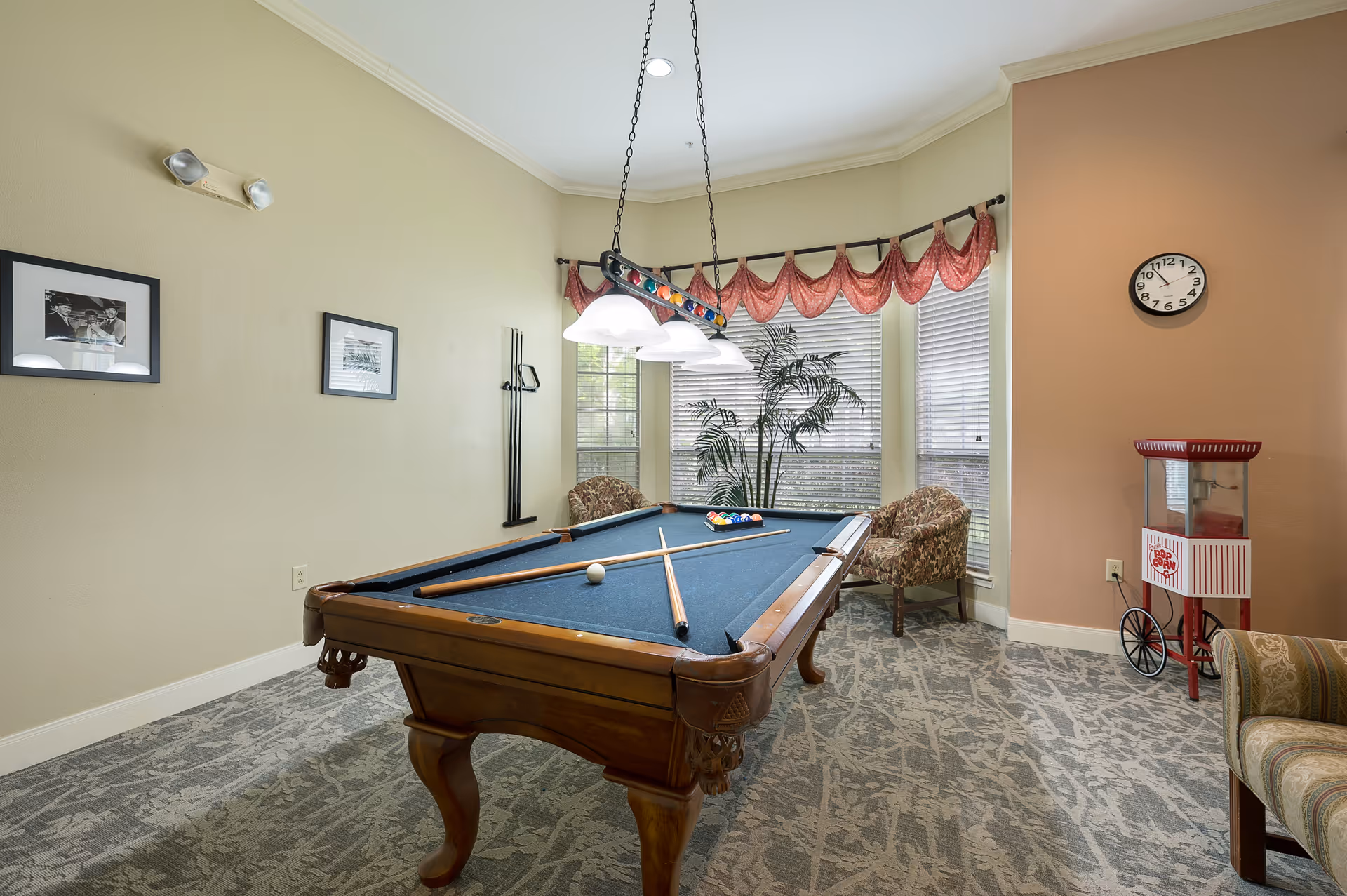 A recreational room with a pool table in the center, two pool cues and a white cue ball on the table. The room has large windows with red valance curtains, two patterned armchairs, a popcorn machine on wheels, a wall clock, and framed black and white photos on the wall. The carpet has a patterned design and the walls are painted in light beige and peach tones.