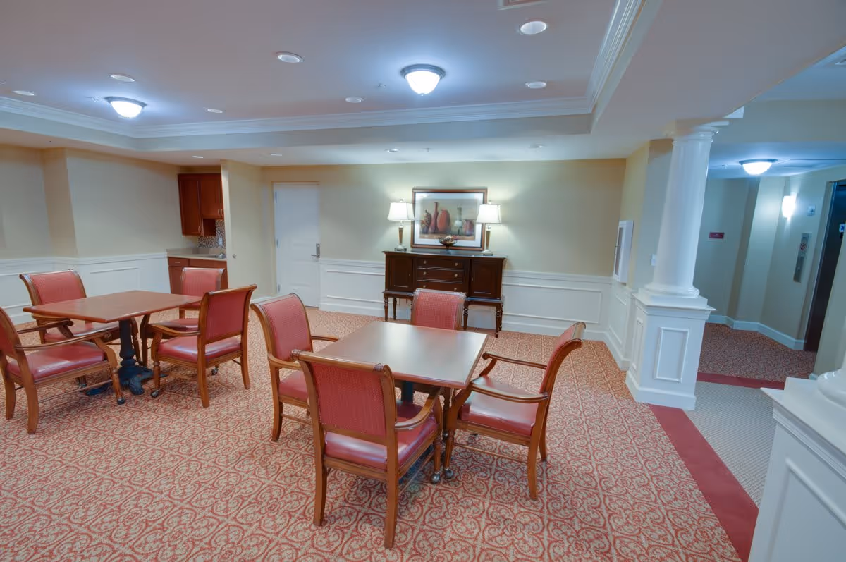 Communal dining/activity room with wooden tables and red upholstered chairs on patterned carpet, a sideboard with lamps and framed art against the far wall.