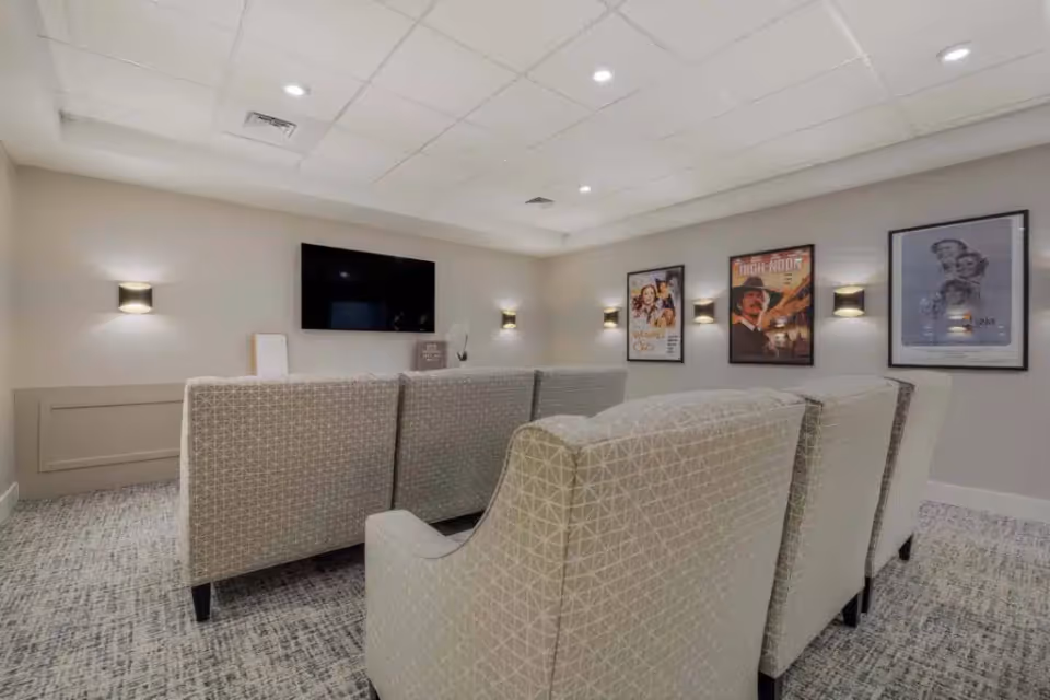 A cozy media room with a wall-mounted flat screen TV, several patterned armchairs arranged in rows facing the TV, soft wall sconces providing ambient lighting, and framed movie posters on the walls.