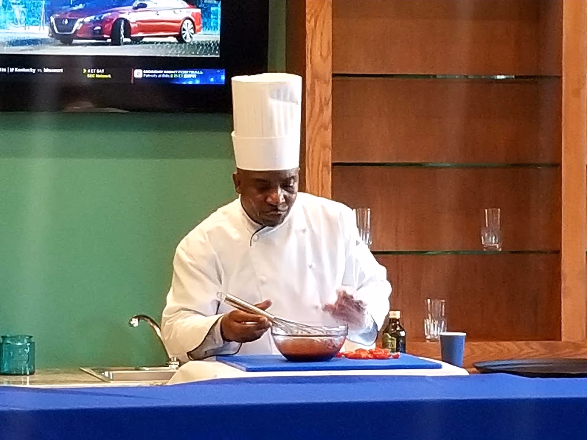 A chef wearing a white uniform and tall white chef hat is whisking a bowl of ingredients on a blue tablecloth-covered table. Behind him is a wooden shelving unit with glass shelves holding a few glasses, and a green wall with a mounted TV screen showing a car advertisement.