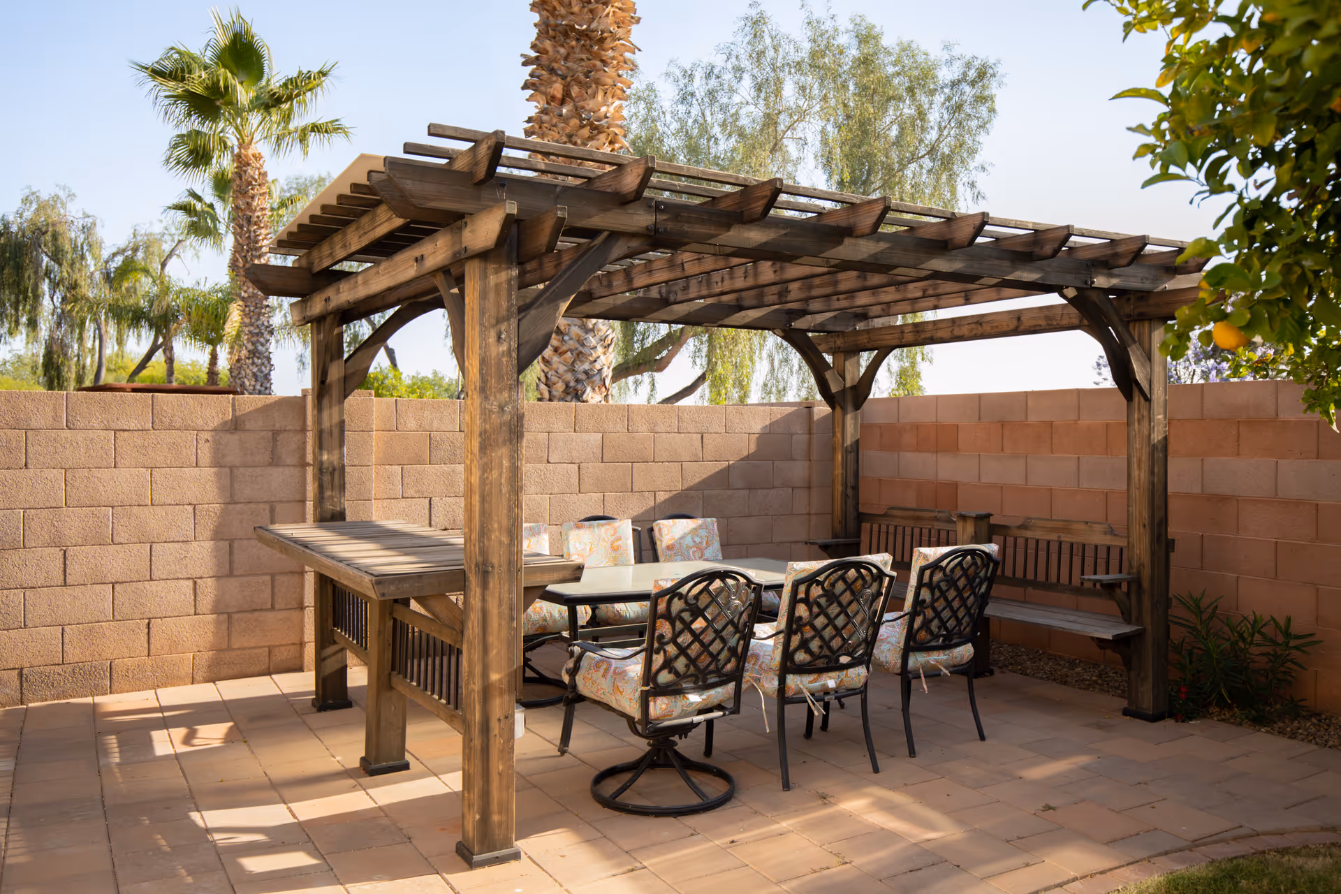 A wooden pergola shades a patio dining set with cushioned chairs and benches against a cinderblock wall with palm trees in the background.