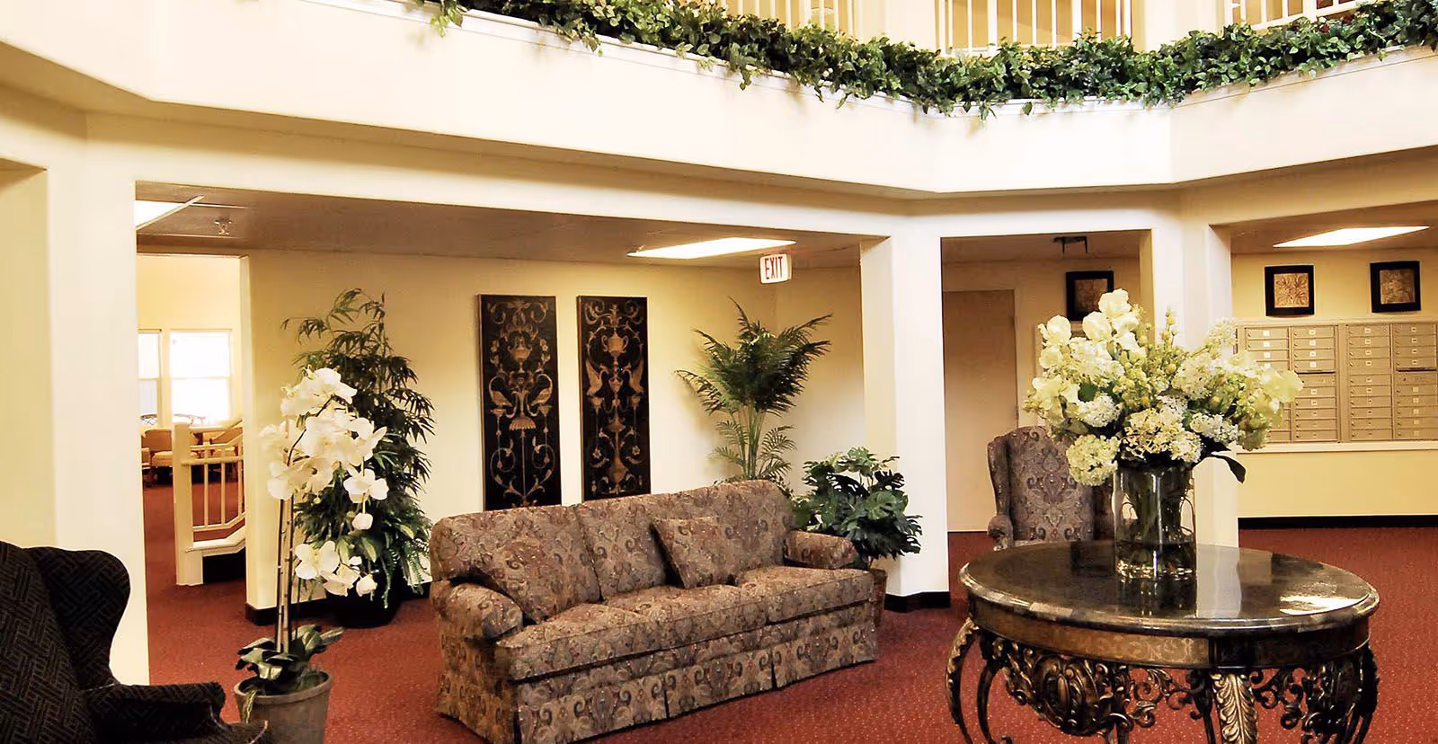 A cozy interior common area with a patterned sofa, armchairs, and a round ornate table with a large vase of white flowers. The space has red carpeting, cream-colored walls, decorative wall art, and several potted plants. There is a mail area with multiple mailboxes visible in the background.