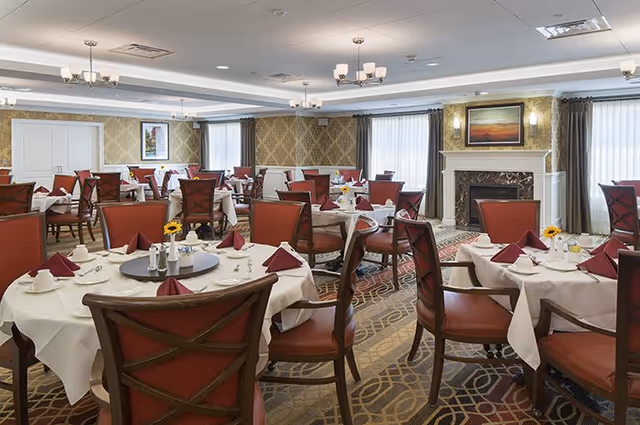 A spacious dining room with multiple round tables covered in white tablecloths, each set with folded burgundy napkins, cups, and small flower arrangements. The room features patterned wallpaper, large windows with curtains, chandeliers, and a fireplace with a framed painting above it.