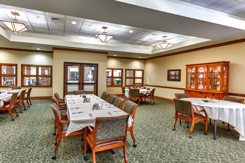 Spacious dining room in a senior living facility with multiple set tables and chairs, carpeted floor, and a display cabinet.