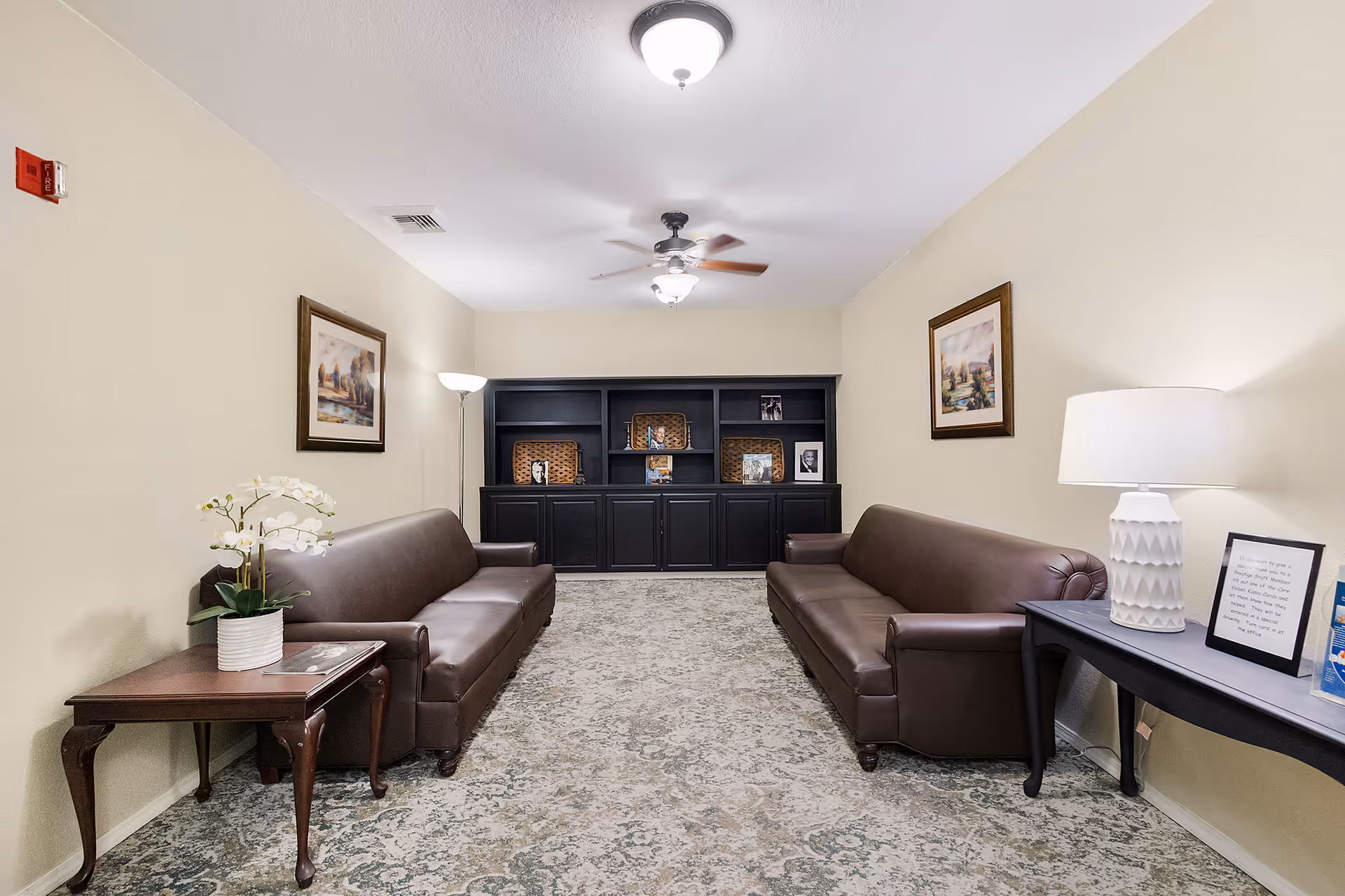 A cozy living room with two brown leather sofas facing each other, a patterned carpet, a black built-in bookshelf with decorative items, two framed paintings on beige walls, a floor lamp, a ceiling fan with light, a side table with a white orchid plant, and a console table with a white lamp and framed text.