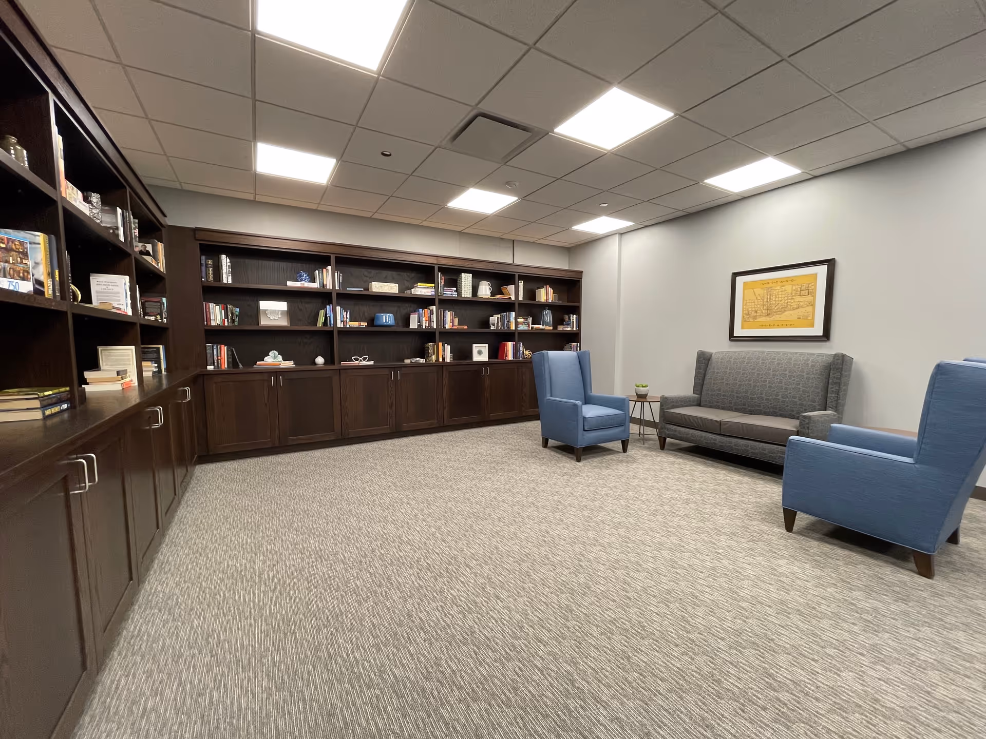 A quiet lounge area with a large dark wood bookshelf filled with books and decorative items along one wall. The room has a gray carpet, light gray walls, and a white ceiling with recessed lighting. There are two blue armchairs and a gray sofa arranged around a small side table with a plant. A framed picture hangs on the wall above the sofa.