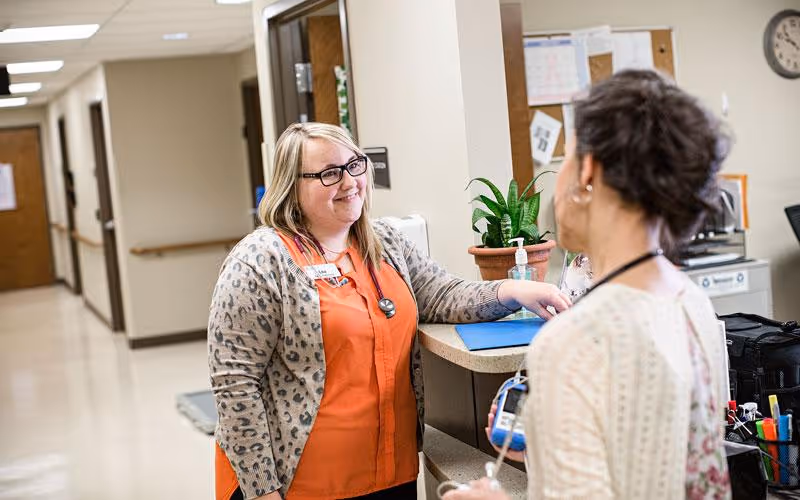 A healthcare worker wearing glasses and an orange shirt with a leopard print cardigan is smiling and talking to another person at a reception desk in a hallway of a senior living facility. The desk has a potted plant, hand sanitizer, and office supplies. The background shows a bulletin board and a clock on the wall.