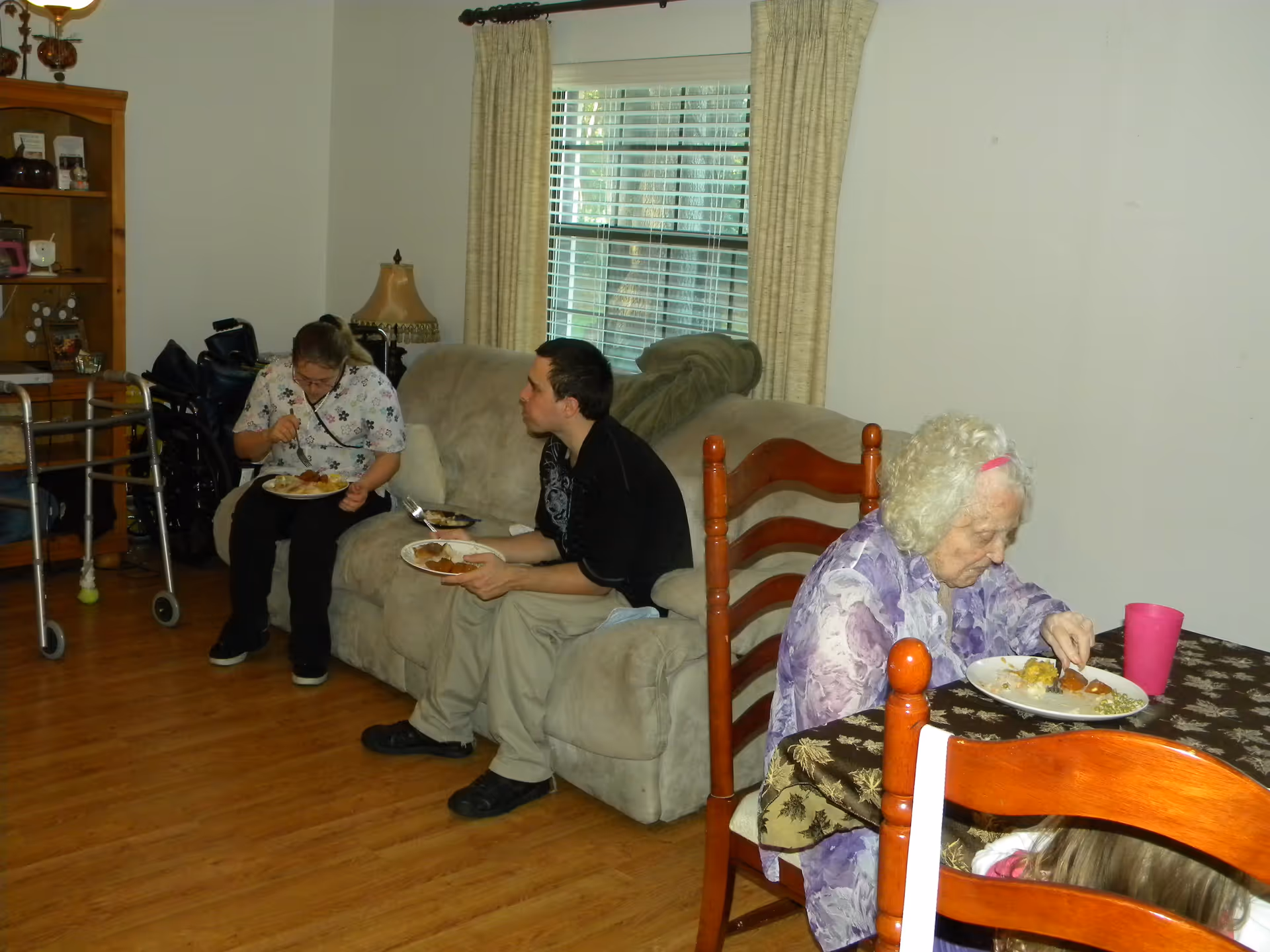 Three people eating in a combined living and dining area—one at a table and two on a couch—near a window with a walker visible nearby.