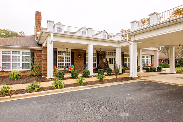 Covered porte-cochere and front entrance of a brick senior living facility with white columns, seating, and landscaped beds.