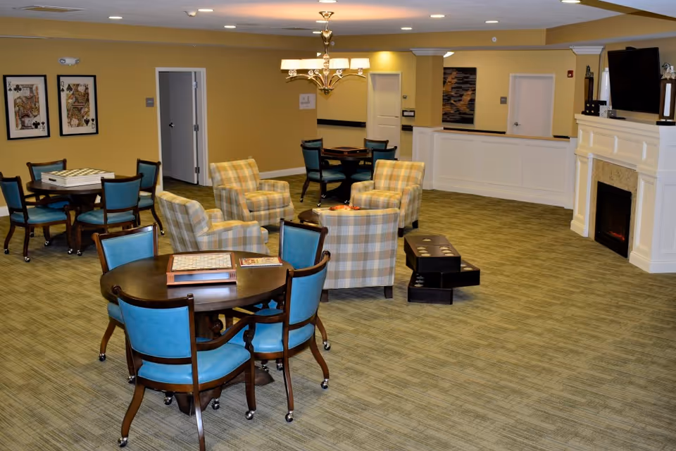 A spacious common area with multiple seating arrangements including blue cushioned chairs around dark wooden tables and plaid upholstered armchairs. The room features a fireplace with a mounted TV above it, framed artwork on the walls, and a chandelier hanging from the ceiling. The carpet is greenish, and the walls are painted yellow. There are also board games on the tables and a set of drawers on the floor.
