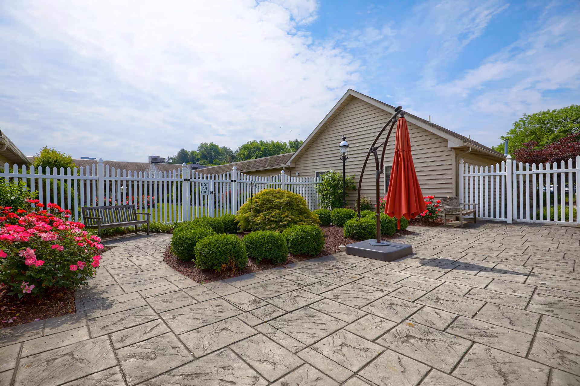 Outdoor patio area with stamped concrete flooring, surrounded by a white picket fence. There are green bushes and pink flowers planted along the edges, two wooden benches, a red umbrella on a stand, and a beige building with a sloped roof in the background under a partly cloudy sky.