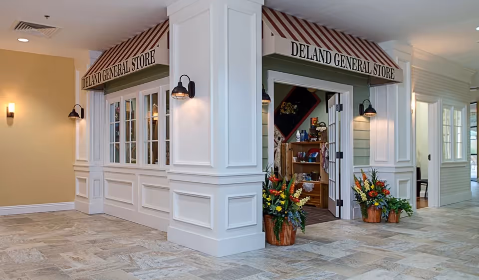 Interior view of a general store named 'Deland General Store' inside a facility. The store has a striped awning with the store name, white paneled walls, and two wall-mounted lights. There are two large flower arrangements in baskets placed on the floor near the entrance. Inside the store, shelves with various items are visible.