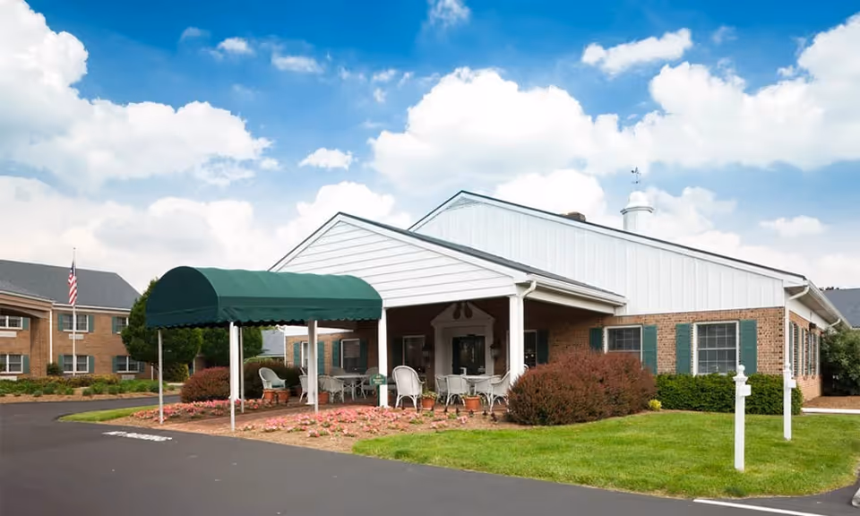 Exterior view of a senior living facility building with a covered entrance featuring a green awning, outdoor seating with white chairs and tables, surrounded by landscaped bushes and flowers under a partly cloudy blue sky.