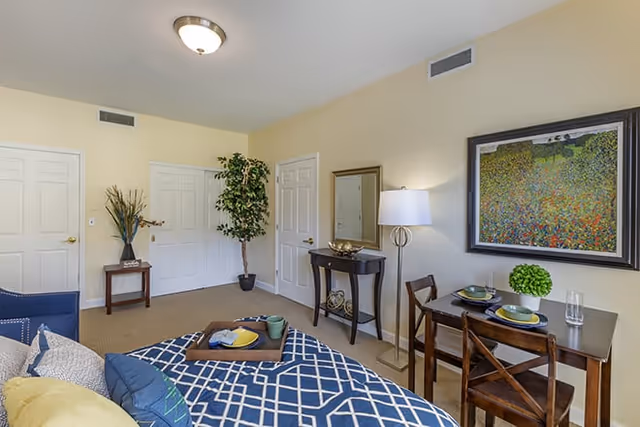 A well-lit bedroom with a bed covered in a blue and white patterned blanket and multiple pillows. There is a small wooden dining table with two place settings and a green plant on top. A floor lamp stands next to a small console table with decorative items and a mirror above it. The walls are light yellow, and there are three white doors and a tall potted plant in the corner. A colorful framed painting hangs on the wall above the dining table.
