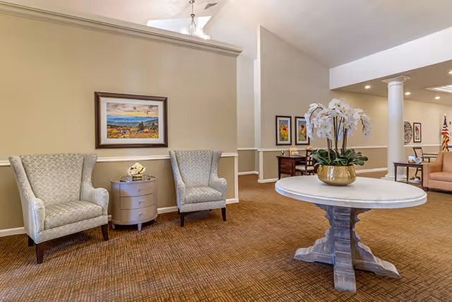 Bright senior living lobby with two upholstered armchairs and side table against a wall and a round center table topped with a floral arrangement.