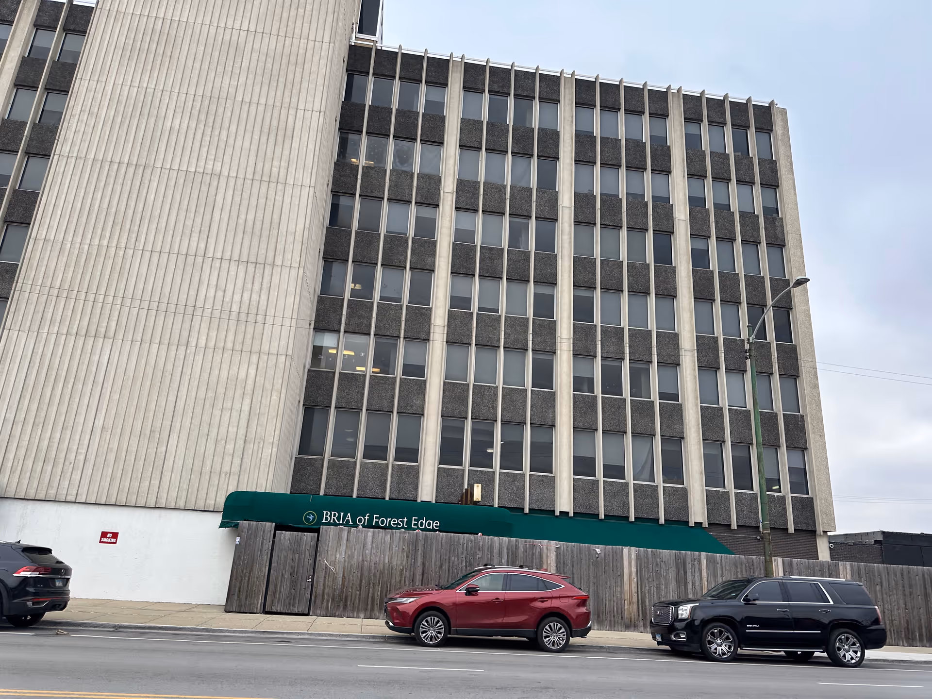 Exterior view of a multi-story building with a green awning that reads 'BRIA of Forest Edge'. Three cars are parked along the street in front of the building, and a wooden fence is visible beneath the awning.