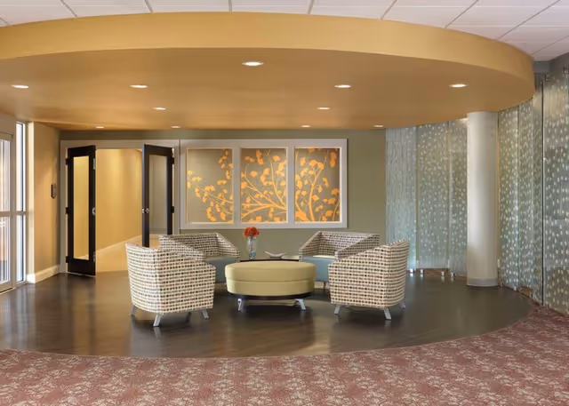 A modern seating area in a senior living facility with four patterned armchairs arranged around a round, light green ottoman. Behind the chairs is a wall with three framed panels featuring a decorative orange leaf design. The space has a curved ceiling feature and a curved glass wall with a dotted pattern on the right side. The floor transitions from a dark wood area under the seating to a patterned carpet in the foreground.