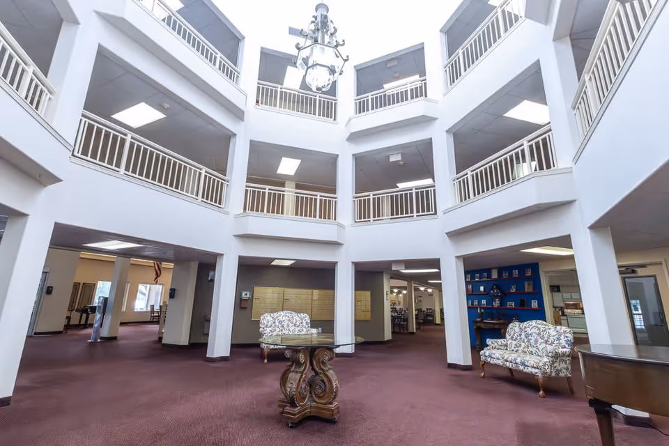 Interior view of a spacious senior living facility lobby with a high ceiling and multiple balconies overlooking the area. The room features a maroon carpet, two floral upholstered chairs, a round glass-top wooden table, and a grand piano. Mailboxes are mounted on the wall, and there is a blue display shelf with various items. The space is well-lit with ceiling lights and natural light from windows.