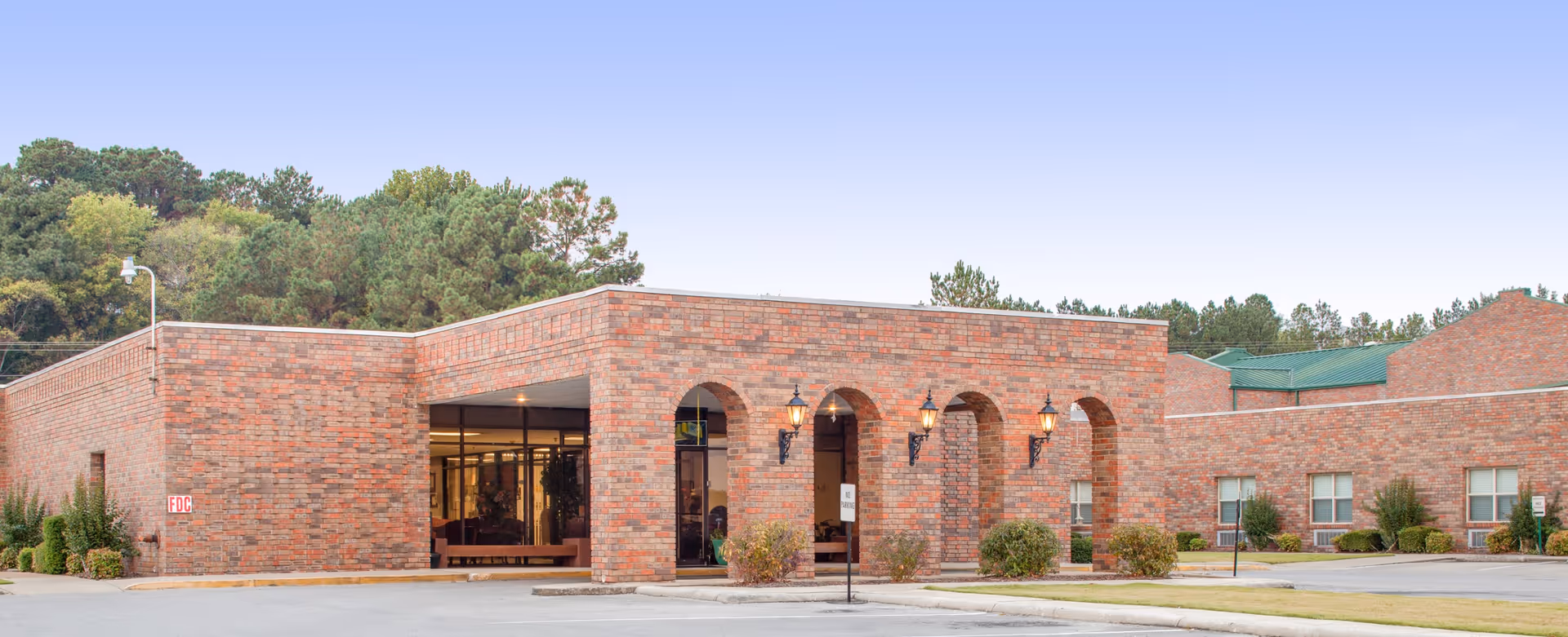 Exterior view of a single-story brick building with arched entryways and outdoor wall lanterns, surrounded by a parking area and greenery with trees in the background under a clear sky.